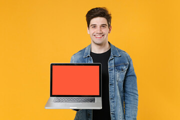 Smiling young man guy in casual denim jacket posing isolated on yellow wall background studio portrait. People lifestyle concept. Mock up copy space. Hold laptop pc computer with blank empty screen.