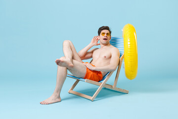 Curious young man in orange shorts glasses sit on deck chair isolated on blue background studio....