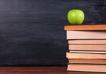 Green apple on big stack of books,  on brown wooden desk with blackboard and chalk streaks in background. Side view, copy space, close-up. Learning, education, knowledge, library, love reading concept