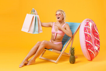 Amazed young girl in pink swimsuit glasses sit on deck chair isolated on yellow background. People summer vacation rest concept. Pointing index finger on package bag with purchases after shopping.