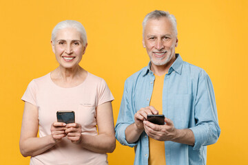 Smiling elderly gray-haired couple woman man in casual clothes isolated on yellow wall background studio portrait. People lifestyle concept. Mock up copy space. Using mobile phone, typing sms message.