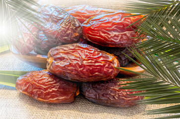 Closeup of date fruits in wooden plate, background of linen textured napkin with leaves of date palm. Top view, closeup, Beams of sunlight from left of frame
