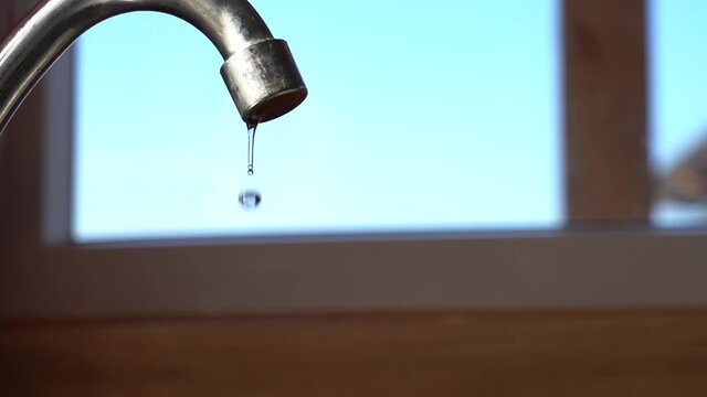Dripping water from a faucet next to a window in the kitchen. Drops of water drip quickly into the water supply. Daylight. Blue sky outside the window.