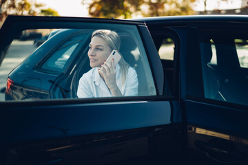 Happy female entrepreneur with open car door making international conversation via modern cellphone gadget, successful business woman using automobile service transportation for getting to destination