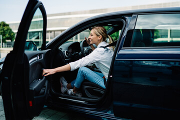 Fototapeta na wymiar Positive hipster girl dressed in casual apparel closing automobile car for journey getaway from city lifestyle, happy cheerful female tourist 20 years old smiling and feeling good from steering drive