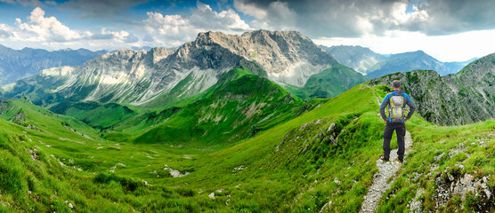 Hiker Man on Hiking Trail with Backpack standing relaxed and enjoying panoramic view to mountains. Alps, Hohe Gaenge, Allgau, Bad Hindelang, Hinterstein, Bavaria, Germany.