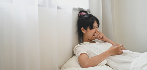 An ill woman holds a glass of water and consuming a pill while lying on the bed in the bedroom.