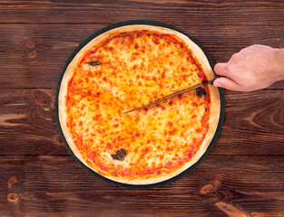 The chef cuts the margherita pizza with a kitchen knife. Delicious pizza with mozzarella, bocconcini and basil leaves on slate round platter which is on wooden background, top view