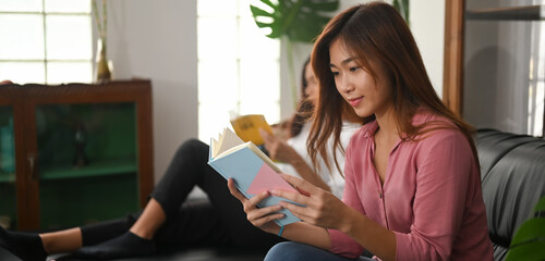 Young Asian women are reading a book while sitting together on a black leather sofa.
