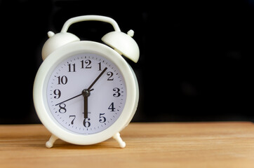 white alarm clock stands on a wooden table against a black wall, copy space