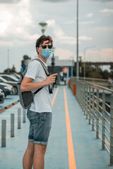 Obraz premium Young man in white t-shirt, jeans shorts, sunglasses, surgical mask and backpack on his back looking back in the parking lot