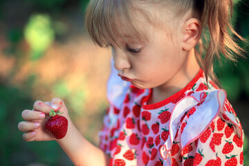 A little girl looks in surprise at a strawberry in her hands. The child makes faces