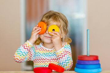 Cute little toddler girl playing alone with colorful wooden rainbow pyramid and toys at home or...