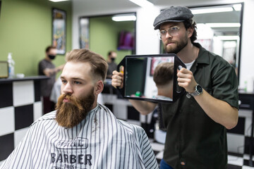 Making haircut look perfect. Young bearded man getting haircut by hairdresser while sitting in chair at barbershop