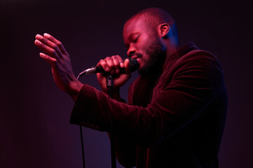 portrait of a dark-skinned handsome guy in dark jacket and t-shirt holds a microphone in his hands and emotionally sings in a dark studio with red and blue light