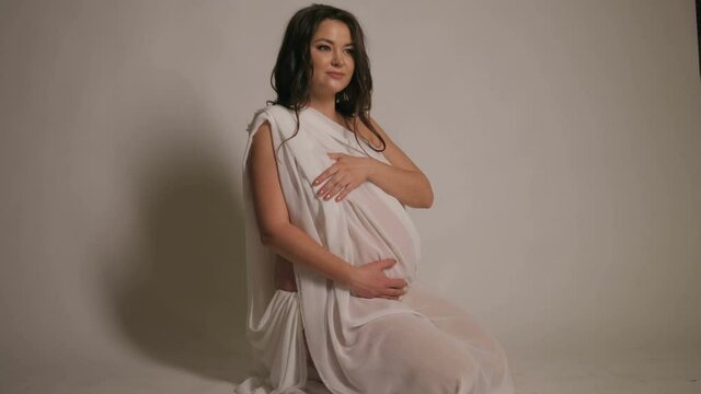 Happy beautiful pregnant woman sitting on the floor in the Studio on a white background wrapped in silk fabric. Naked pregnant girl posing in the Studio. Slow motion.