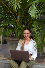 Freelance. Woman Working On Laptop Near Tropical Plants. Young Woman With Computer Wearing Casual Clothes And Enjoying Summer Vacation. Beautiful Model In Glasses Against Palm Leaves.