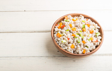 Fototapeta na wymiar Pearl barley porridge with vegetables in wooden bowl on a white wooden background. Side view, copy space.