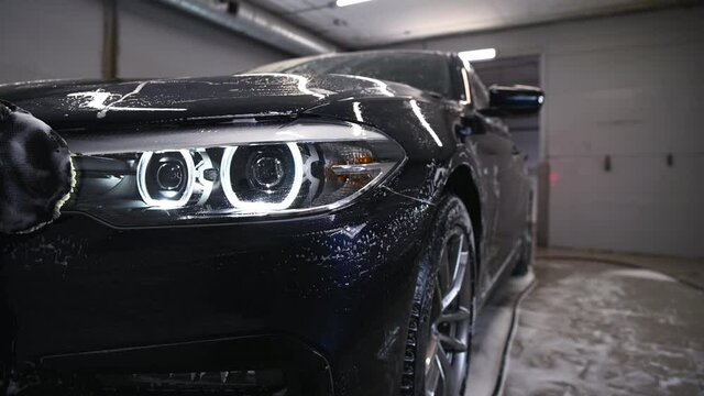 Car detailing - the man washes headlights of a car with a hand covered with a special wash glove, close-up.