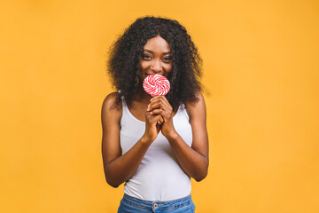Fototapeta na wymiar Smiling american african girl holding candy lollipop. Young cheerful woman with lollipop isolated over yellow background, copy space. Sweet life and confectionary.