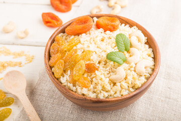 Bulgur porridge with dried apricots,  raisins and cashew in wooden bowl on a white wooden background and linen textile. Side view, selective focus.