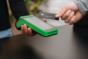 Closeup of male right hand holding bank credit card on a NFC payment terminal held by a woman's...