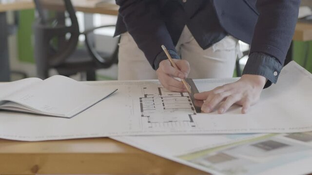 A young male architect is studying a building plan, measures the dimensions of the blueprint with a ruler and a pencil on the desktop in the design office.
