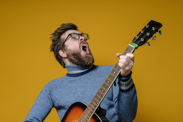 Hipster in an old sweater plays an acoustic guitar and sings loudly, on a yellow background. Hobbies, lifestyle.