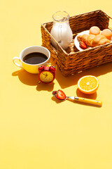 Wicker basket with croissant, milk, a cup of morning coffee and an alarm clock. Breakfast on a yellow background