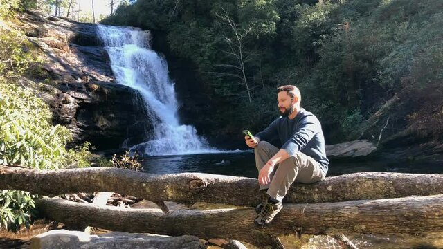 504 Hiker relaxing on the phone on a log in a river with a waterfall behind him
