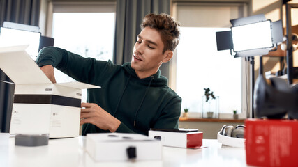 Fototapeta premium Time for Unpacking. Young male technology blogger recording video blog or vlog about new camera lens and other gadgets at home studio. Blogging, Work from Home concept
