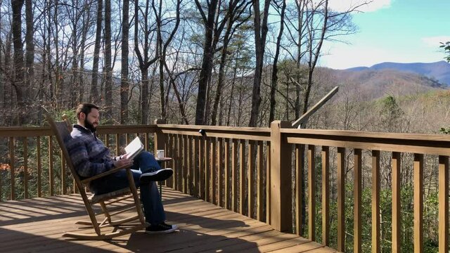 508 Man relaxing in a cabin in the woods while reading a book and drinking coffee in a rocking chair