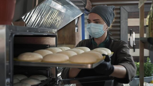 small business, young cute female baker in medical mask puts bake with slices of dough for baking in an electric furnace while working in bakery after removing quarantine