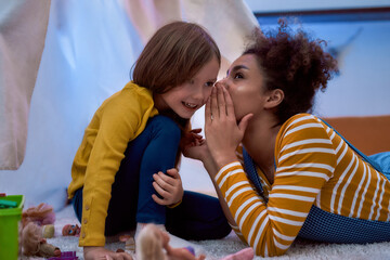 African american woman baby sitter entertaining caucasian cute little girl. They are gossiping and telling secrets sitting in kids room
