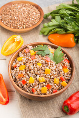 Buckwheat porridge with vegetables in wooden bowl on a white wooden background and linen textile. Side view, close up, selective focus.