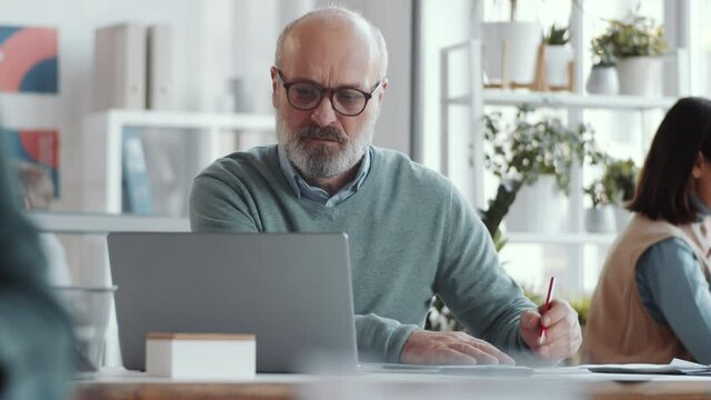 Senior businessman in glasses and smart casual outfit sitting at desk, typing on laptop and writing in notepad while working in open space office