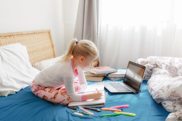 Caucasian girl child sitting in bed and learning online on laptop Internet. Virtual class lesson school on video chat during self-isolation quarantine at home. Distant remote video education class.