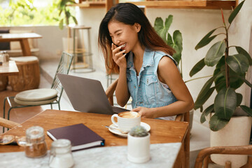 Woman Chatting Online At Cafe. Asian Girl In Jeans Clothes Using Laptop For Online Meeting. Comfortable Digital Nomad Lifestyle With Modern Technologies For Communication.