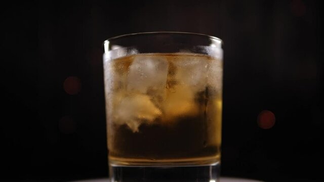 Close-up of a whiskey glass with ice spinning on a black background with a flashing garland. Slow motion.