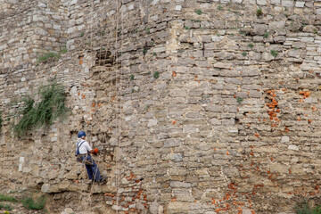 Fototapeta na wymiar A worker on ropes repairs an old stone wall, restoration of medieval structures and landmarks. High-altitude works.