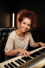 Beautiful girl happily looking in camera playing on electric piano in studio. Attractive musician working in recording studio