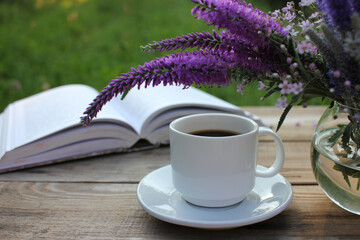 Cup of coffee, open book and bunch of purple summer flowers in glass vase on wooden table in the...