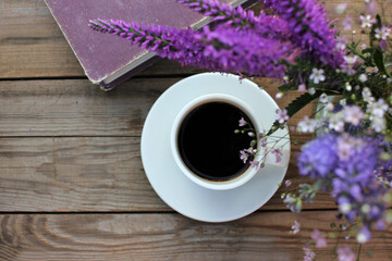 Cup of coffee, book and bunch of purple summer flowers in glass vase on wooden table in the garden....