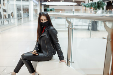 Girl with medical black mask and mobile phone in a shopping center. Coronavirus pandemic. A woman with a mask is standing in a shopping center. A girl in a protective mask is shopping at the mall