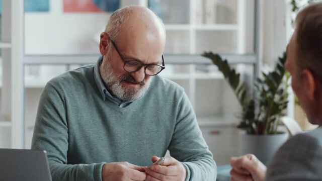 Tracking shot of cheerful senior businessman talking with male colleague and taking notes while sitting at desk in the office