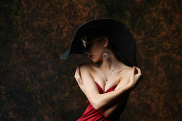 beautiful girl in a wide-brimmed hat and in a burgundy evening dress hugs herself on a black background