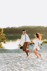 Positive couple running on the sand, sunset, stylish couple with a suitcase and flowers in a basket.