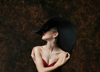 beautiful girl in a wide-brimmed hat and in a burgundy evening dress hugs herself on a black background