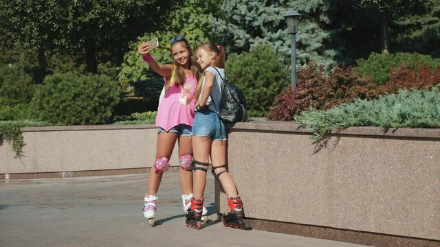 Two modern smiling teenage girls roller skating in a city park stopped to take a selfie