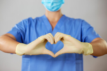 Female doctor in a face mask and gloves forming a heart in front of blue shirt with her fingers. Medical worker makes a positive symbol with fingers in latex gloves.